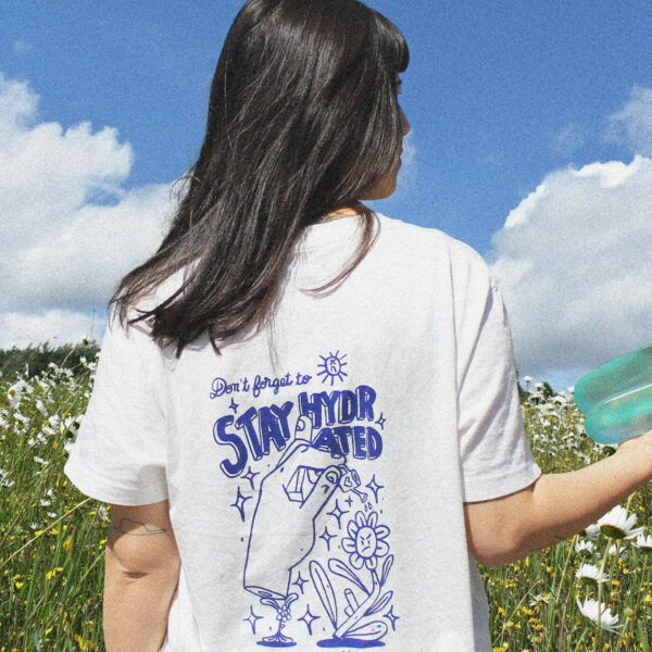 Femme portant le t-shirt blanc sérigraphié bleu, de dos. Elle tient une gourde dans sa main. Sur fond de ciel bleu et prairie fleurie. Le dessin du teeshirt représente une petite fleur qui porte une main qui elle même tient un arrosoir et qui arrose une grande fleur boudeuse. Avec le message "Stay hydrated"