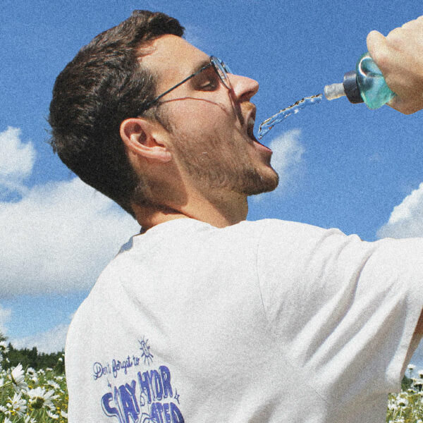 Homme portant le t-shirt blanc sérigraphié bleu, de profil. De l'eau part d'une gourde en direction de sa bouche. Sur fond de ciel bleu et prairie fleurie